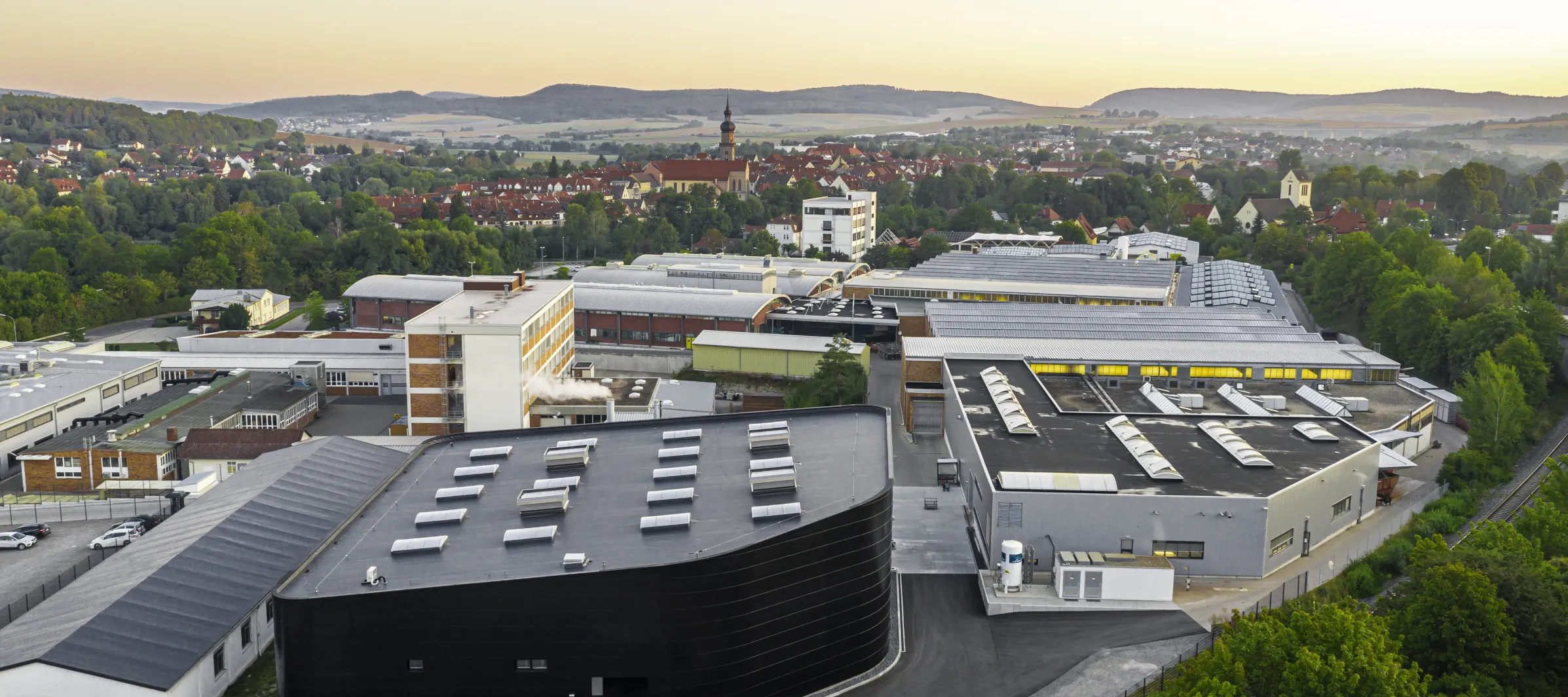 Eine Luftaufnahme bei Sonnenuntergang mit dem Produktionsgelaende der Reich GmbH im Vordergrund und Mellrichstadt und Bergen im Hintergrund 
